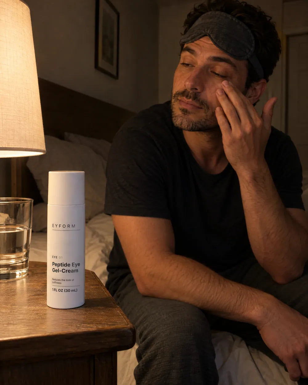 A man on the edge of his bed at night with a sleep mask pushed up, patting EYE 01 under his eye